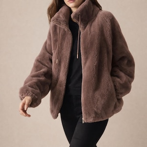 May include: A brown faux fur jacket with a high collar and a zip fastening. The jacket has long sleeves and a soft, textured appearance. The model is wearing black trousers and a black top.