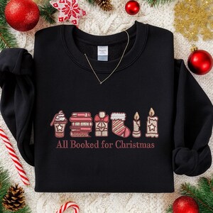 May include: Black sweatshirt featuring embroidered Christmas motifs: a coffee cup, books, a gift, a stocking, and candles. The text "All Booked for Christmas" is embroidered below. A gold necklace is visible.