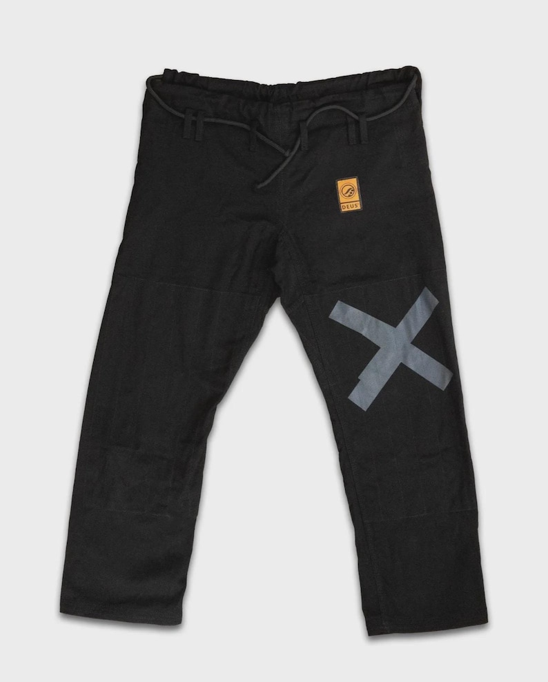 May include: Black martial arts trousers with a grey X graphic on the left leg. The trousers have a drawstring waist and a small, square patch with text. The trousers are made of a durable material.