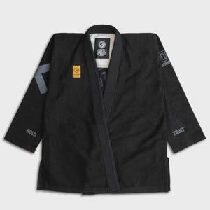 May include: Black martial arts gi with the brand name "DEUS" on the chest and sleeve. The gi has a V-neck design and features grey accents on the sleeves, with the words "HOLD" and "TIGHT" printed on the sleeves.