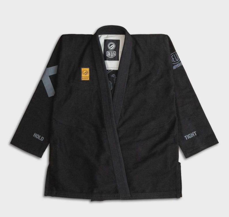 May include: Black martial arts gi with the brand name "DEUS" on the chest and sleeve. The gi has a V-neck design and features grey accents on the sleeves, with the words "HOLD" and "TIGHT" printed on the sleeves.