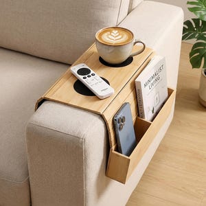 May include: A bamboo sofa arm tray with a built-in cup holder, remote control holder, and storage compartment. A cup of coffee, a remote, a phone, and a book titled "Minimalist Living" are on the tray. The tray is on a beige sofa.