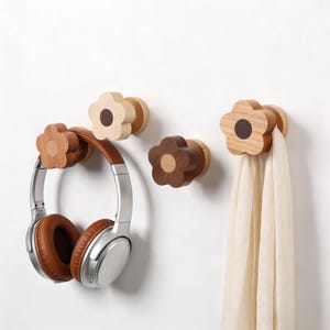 Op de afbeelding: Houten bloemvormige wandhaken in verschillende bruin- en beigetinten. Eén haak houdt een bruine en zilveren koptelefoon vast, een andere ondersteunt een crèmekleurige sjaal. De haken zijn op een witte muur gemonteerd.