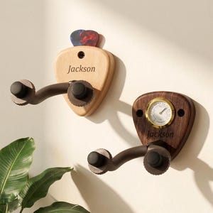 Op de afbeelding: Twee gitaarvormige wandhouders. Eén is van licht hout met de naam "Jackson" en plectrums. De andere is van donker hout met een thermometer en de naam "Jackson". Beide hebben een bruine arm om een gitaar vast te houden.