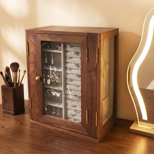 May include: A dark wooden jewellery cabinet with a glass door, displaying rings, earrings, and necklaces. The cabinet has a dark brown finish with brass fittings. A makeup brush holder is on the left.