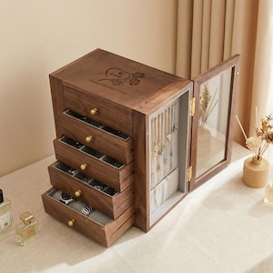 May include: A wooden jewellery box with multiple drawers and a glass-door compartment. The box is a warm brown colour with gold-coloured knobs and a decorative engraving on the top. The drawers are open, revealing jewellery and watches.