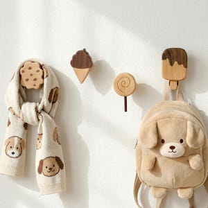 Puede incluir: Bufanda crema con estampado de cachorros colgada junto a ganchos de madera con forma de galleta, cono de helado, piruleta y polo. Una mochila marrón claro con forma de cachorro cuelga del gancho del polo.