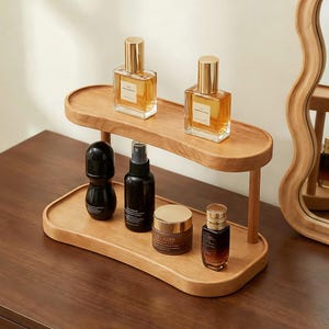 Peut inclure: Un organisateur de vanit&eacute; en bois &agrave; deux niveaux avec un design incurv&eacute;. L'&eacute;tag&egrave;re sup&eacute;rieure contient deux flacons de parfum avec des bouchons dor&eacute;s. L'&eacute;tag&egrave;re inf&eacute;rieure affiche un d&eacute;odorant noir, un vaporisateur et deux pots cosm&eacute;tiques. L'organisateur est sur une surface en bois.