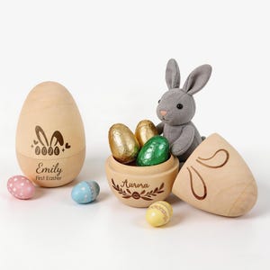 Op de afbeelding: Houten paasei met konijnenoren en de naam "Emily" en "First Easter" erop gedrukt. Een grijze pluche konijn zit naast een houten kom gevuld met in folie verpakte chocolade-eieren. Decoratieve paaseieren zijn verspreid.