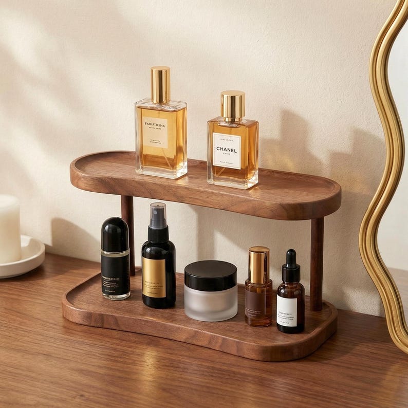 May include: A two-tiered, dark wood vanity organizer holding various beauty products. Two perfume bottles sit on the top shelf, while the lower shelf displays a black rollerball, spray bottle, jar, and two smaller bottles. A gold-framed mirror is visible.
