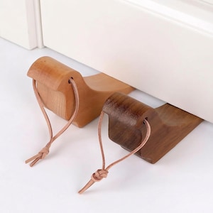 May include: Two wooden doorstops in light and dark brown shades. Each doorstop features a curved design and a leather loop for hanging. The doorstops are made of wood and are designed to prevent doors from closing.