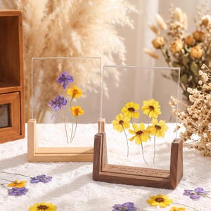 May include: Two clear acrylic frames with pressed flowers, one with purple and yellow blooms, the other with yellow flowers. The frames are set in wooden stands, one light and one dark, with a quote on the dark stand.