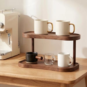 Peut inclure: Un pr&eacute;sentoir en bois &agrave; deux niveaux pr&eacute;sente des tasses et des mugs &agrave; caf&eacute;. Deux mugs cr&egrave;me avec des poign&eacute;es dor&eacute;es sont sur le niveau sup&eacute;rieur. Le niveau inf&eacute;rieur contient un mug noir avec une poign&eacute;e en bois, une tasse en verre transparent et un mug blanc.