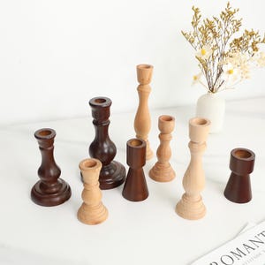 Puede incluir: Una colección de candelabros de madera de varias formas y tamaños. Los candelabros están hechos de diferentes tipos de madera, incluyendo tonos marrones claros y oscuros. Algunos tienen un diseño torneado clásico, mientras que otros son más modernos. Un jarrón blanco con flores está en el fondo.
