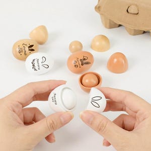 May include: A collection of wooden Easter eggs in various shades of brown and white. Some eggs are decorated with bunny ears and floral designs. One egg is split open, revealing a white interior.
