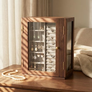 May include: A wooden jewellery cabinet with a glass door displays earrings and rings. The cabinet is brown with a gold knob and hinges. A pearl necklace rests on the wooden surface in front of the cabinet.