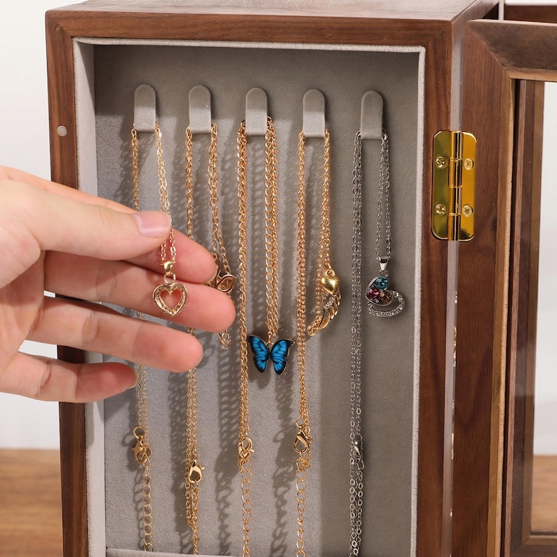 May include: A wooden jewelry box with a glass door displays several necklaces. The necklaces have gold and silver chains with various pendants, including a heart, a butterfly, and a pendant with colorful stones. The box has a grey interior.
