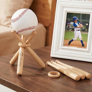 May include: A baseball displayed on a wooden stand with the name "David" engraved on one leg. A white framed photo of a baseball player is next to the baseball. Three miniature baseball bats and a wooden ring are also present.