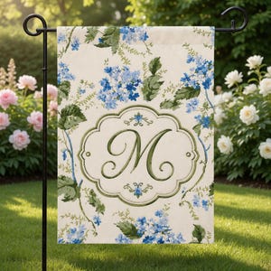 May include: A garden flag with a floral design featuring blue flowers and green leaves on a cream background. The flag has a green framed monogram letter "M" in the center. The flag is displayed on a black metal stand in a garden setting.