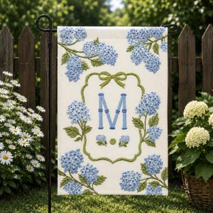 May include: A cream-colored garden flag featuring blue hydrangea blooms and green leaves. The flag has a green bow and a decorative frame around the letter "M" in blue. The flag is displayed on a black metal stand.
