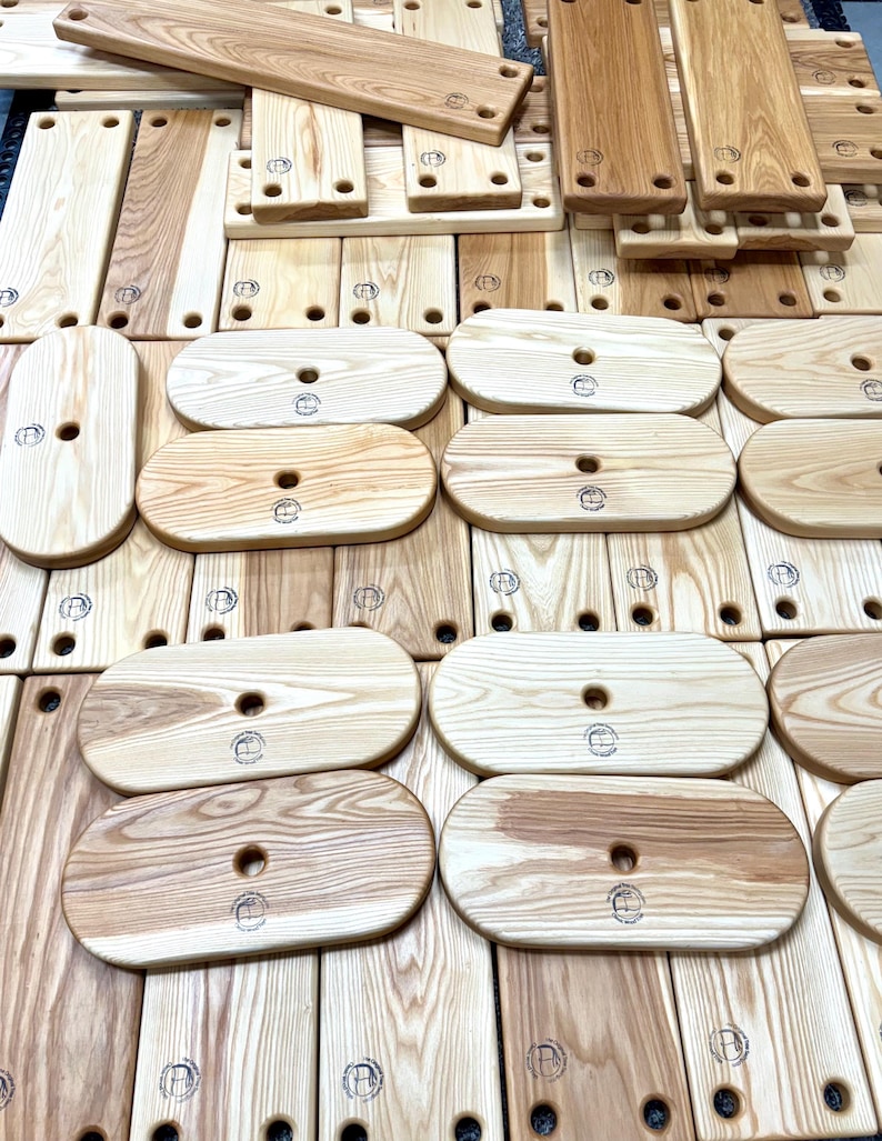 Puede incluir: Una colecci&oacute;n de tablas de cortar ovaladas de madera con un agujero en el centro. Las tablas son de madera clara con un patr&oacute;n de veta natural. Cada tabla tiene un logotipo circular quemado en la superficie.