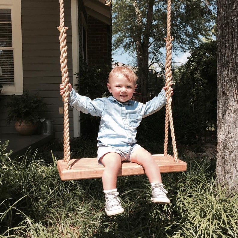 Puede incluir: Un ni&ntilde;o peque&ntilde;o con una camisa de botones azul claro y zapatillas grises est&aacute; sentado en un columpio de madera. El columpio est&aacute; hecho de cuerda natural y tiene un asiento de madera. El ni&ntilde;o sonr&iacute;e y mira a la c&aacute;mara.
