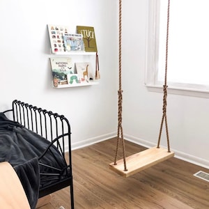 Puede incluir: Un columpio de madera con cuerda natural colgando del techo en una habitaci&oacute;n con paredes blancas y una cama de metal negro. El columpio est&aacute; hecho de una sola pieza de madera y tiene unos 45 cm de ancho.
