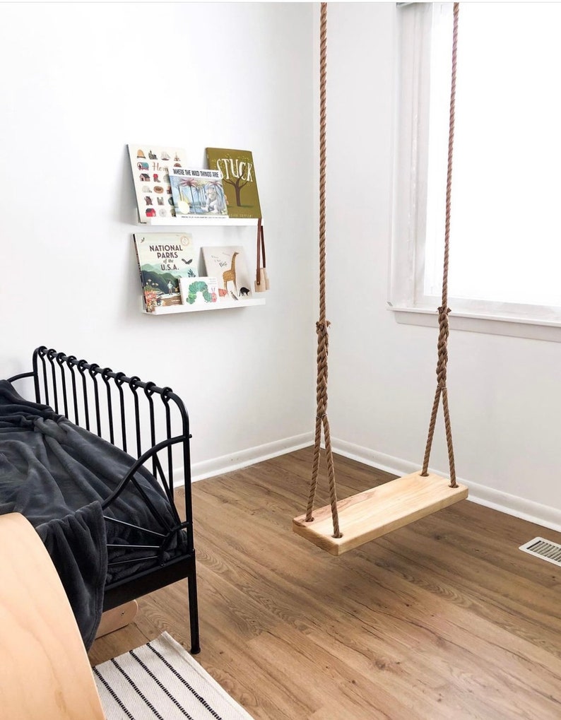 Puede incluir: Un columpio de madera con cuerda natural colgando del techo en una habitaci&oacute;n con paredes blancas y una cama de metal negro. El columpio est&aacute; hecho de una sola pieza de madera y tiene unos 45 cm de ancho.