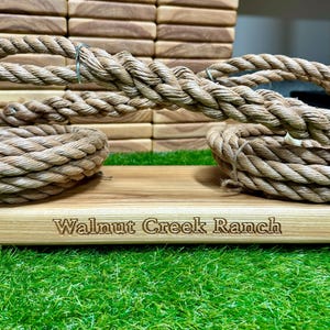 May include: Close-up of a wooden board with the engraved text "Walnut Creek Ranch." Thick, coiled rope rests on the board, with more rope in the background. The rope is a natural beige color, and the board is light brown.