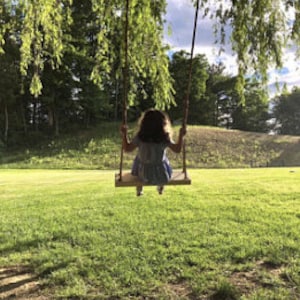 Puede incluir: Una persona joven se sienta en un columpio de madera con un fondo de hierba verde y &aacute;rboles. El columpio est&aacute; hecho de cuerda y tiene un asiento de madera.