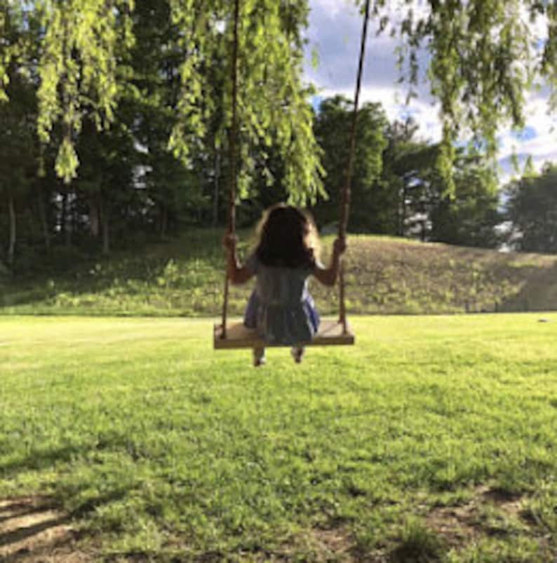 Puede incluir: Una persona joven se sienta en un columpio de madera con un fondo de hierba verde y &aacute;rboles. El columpio est&aacute; hecho de cuerda y tiene un asiento de madera.