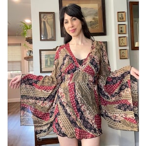 May include: A patterned dress with a V-neck and long, flared sleeves. The dress features a mix of red, black, and tan colors in a patchwork design. The dress has a black trim along the neckline and hem.