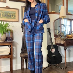 May include: A blue plaid suit with a jacket and matching pants. The jacket has pockets and buttons. The suit is worn with a navy blue top and black boots. The suit's plaid pattern includes blue, white, and burgundy lines.