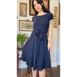 May include: A solid navy blue knee-length dress featuring short sleeves, a round neckline, and a matching fabric tie belt at the waist. The skirt has a flared silhouette with a soft, flowing drape, creating a simple and classic look for any occasion.