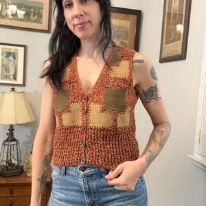 Puede incluir: Un chaleco sin mangas con botones en tonos naranja y marrón. El chaleco tiene cuello en V y parches cuadrados de material similar al ante beige. La prenda se lleva sobre unos vaqueros azules.