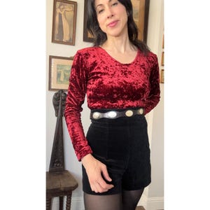 May include: A long-sleeved, deep red crushed velvet top with a round neckline paired with high-waisted black velvet shorts. The outfit is accented by a black leather belt featuring evenly spaced, circular silver-toned metal concho embellishments.