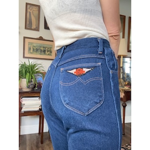 Puede incluir: Vaqueros de mezclilla azul de talle alto con un bolsillo trasero que presenta un parche de Harley-Davidson. El parche tiene forma de alas con texto en naranja y negro. Los vaqueros tienen una construcción clásica de cinco bolsillos.