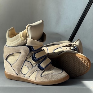 May include: A pair of beige high-top wedge sneakers featuring suede panels, dark blue trim, and three hook-and-loop closure straps across the front. The shoes have a padded collar, a high tongue, and a flat gum rubber outsole with a textured tread.