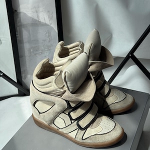 May include: A pair of high-top wedge sneakers in a light beige suede and leather finish. The shoes feature a prominent padded tongue, multiple black-trimmed hook-and-loop velcro straps across the midfoot, and a gum rubber outsole.