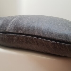 May include: A gray leather pillow with a zipper closure. The pillow has a distressed look and a subtle texture.