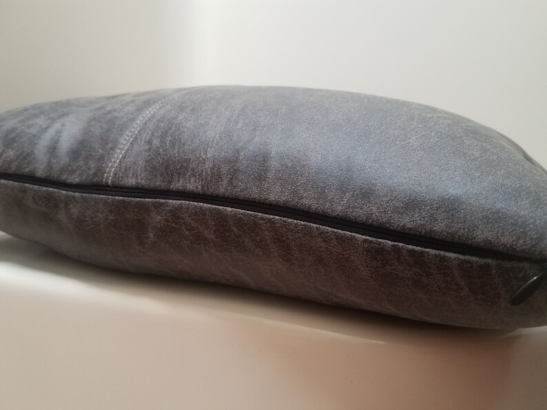 May include: A gray leather pillow with a zipper closure. The pillow has a distressed look and a subtle texture.