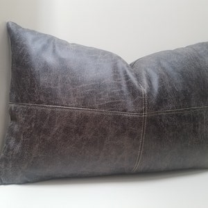May include: A gray leather pillow with white stitching. The pillow has a distressed look and is rectangular in shape.