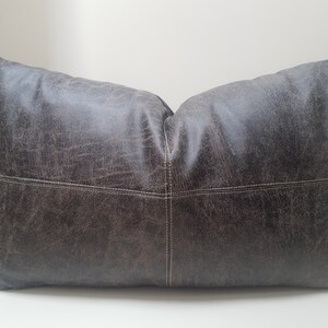 May include: A large, rectangular pillow with a dark gray, distressed leather-like fabric. The pillow has white stitching around the edges and a subtle diamond pattern.