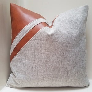 May include: A beige pillow with a brown leather accent. The leather is stitched in a diagonal pattern across the pillow.