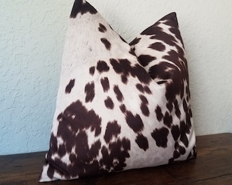 May include: A brown and white cowhide patterned throw pillow. The pillow is made of a soft, plush fabric and has a square shape.