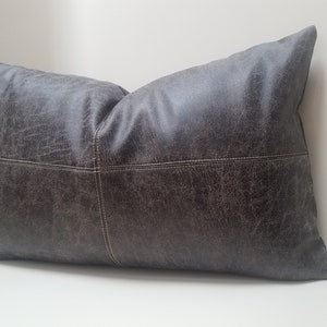 May include: A dark gray rectangular pillow with a textured faux leather cover. The pillow has white stitching along the seams.