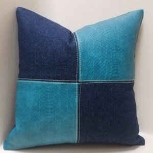 May include: A square pillow with a patchwork design. The pillow is made of two different fabrics: a dark blue denim and a turquoise blue fabric with a textured pattern. The fabrics are sewn together in a checkered pattern with white stitching.