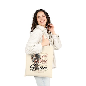 Puede incluir: Un bolso de tela color crema con un estampado gráfico de un lazo con patrones de la bandera estadounidense. El texto dice: Where the Spirit of the Lord is, there is Freedom, junto con la cita 2 Corinthians 3:17 en letra decorativa.
