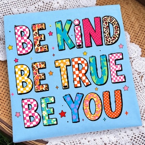 Könnte beinhalten: Ein hellblaues T-Shirt mit dem Text BE KIND BE TRUE BE YOU in großen, bunten Buchstaben. Die Buchstaben zeigen verschiedene Muster wie rosa-weiß karierte Flächen, Leopardenmuster, Blumenmotive und Punkte, akzentuiert durch kleine Sterne.