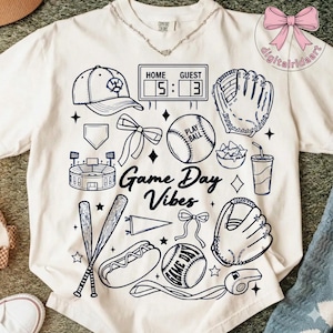 Könnte beinhalten: Cremefarbenes T-Shirt mit Baseball-Motiv. Das Design umfasst eine Baseballkappe, Handschuhe, Basebälle, ein Stadion und den Text "Game Day Vibes". Der Spielstand ist 5-3. Ein lustiges Shirt für Baseballfans.